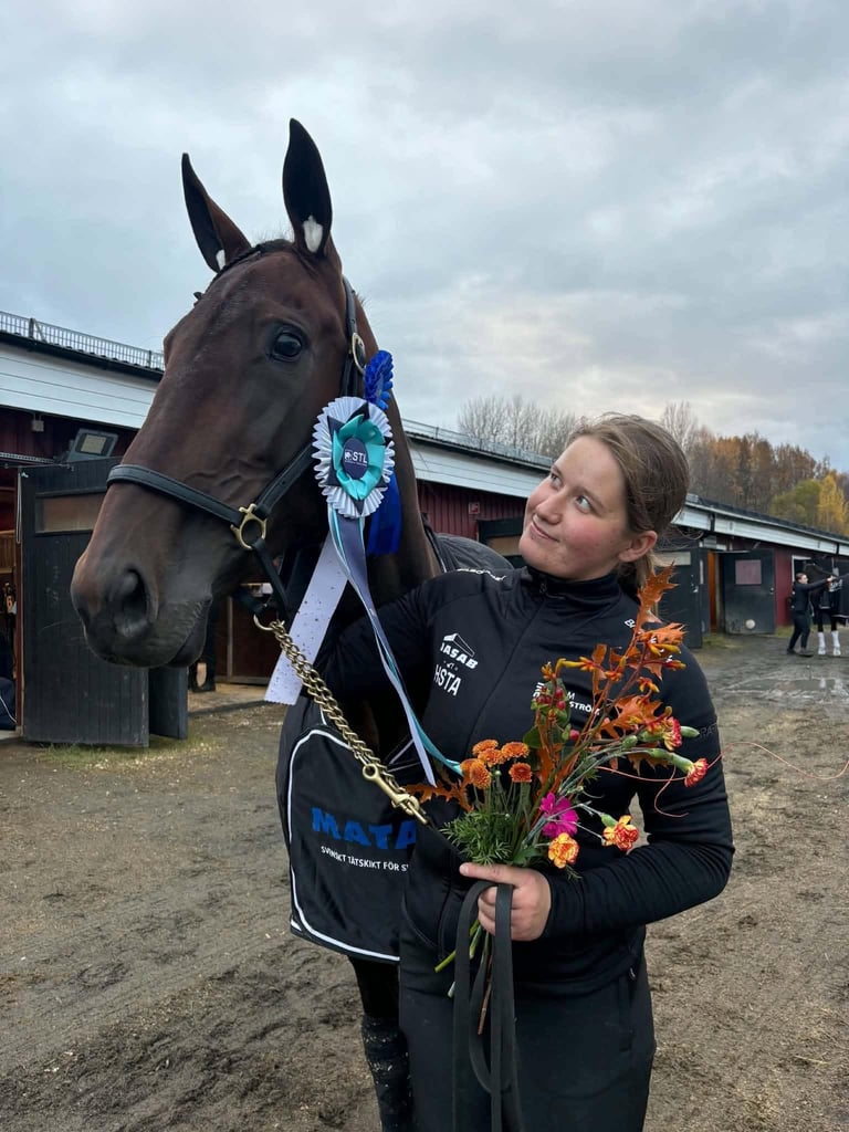Woman in black jacket holding colorful flowers standing next to a brown horse wearing a blue award ribbon at an equestrian facility