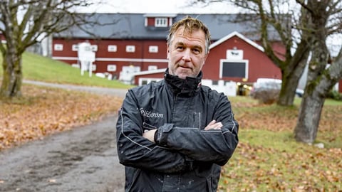 Middle-aged man in dark jacket standing with arms crossed on rural driveway with red barn in background
