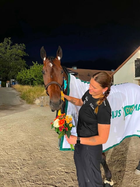 Woman in black shirt poses with a brown horse wearing a decorative flower garland at night