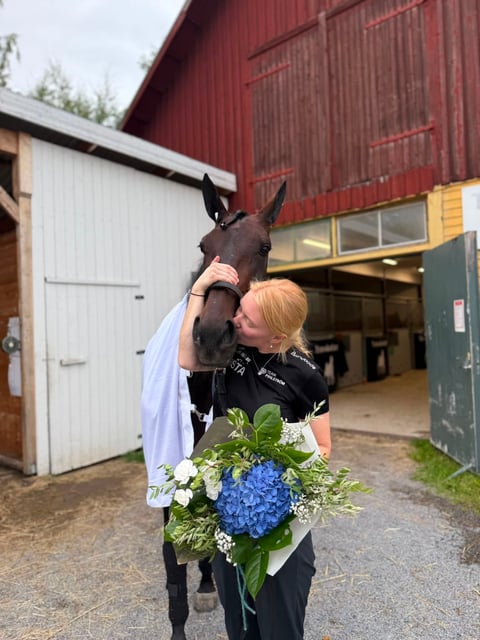 Woman in black jacket holding blue and white flower bouquet while posing with dark brown horse outside red barn