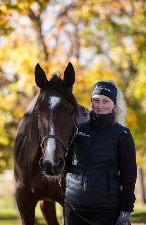 Woman in black jacket standing beside dark brown horse with white blaze on face, autumn foliage background