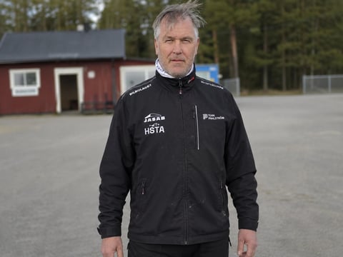 Middle-aged man in black athletic jacket standing outdoors with red barn and trees in background