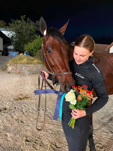 Woman in black jacket holding award bouquet next to brown horse at night at equestrian facility