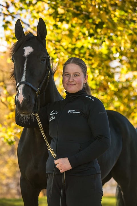 Young woman in black equestrian attire holds a black horse with a white blaze against a blurred yellow foliage background