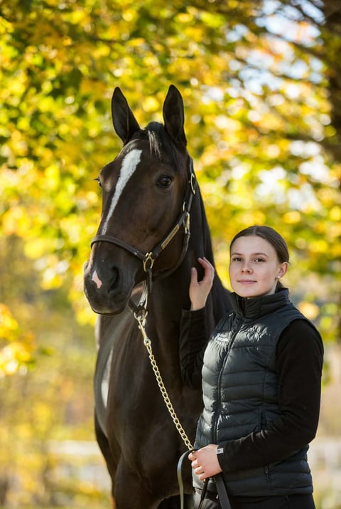 Young girl in black jacket holding halter of dark brown horse with white blaze against yellow autumn foliage