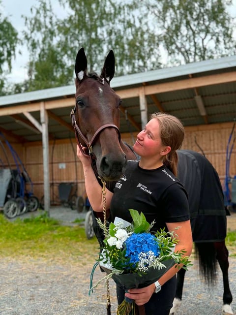 Woman in black shirt holding a horses lead and bouquet of flowers in front of a stable