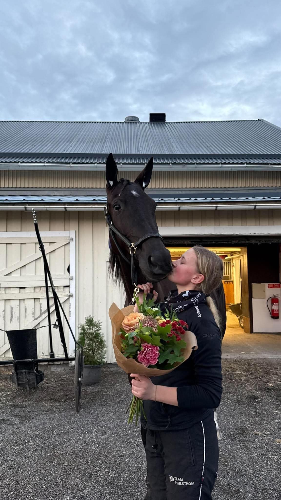 Woman in black clothing kissing a black horse while holding a colorful flower bouquet at a stable