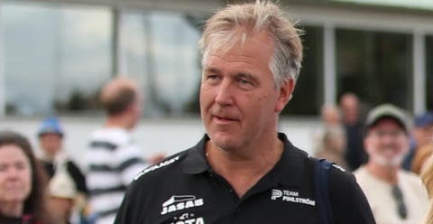 Middle-aged man with gray hair wearing a black racing team shirt, speaking outdoors with spectators in the background