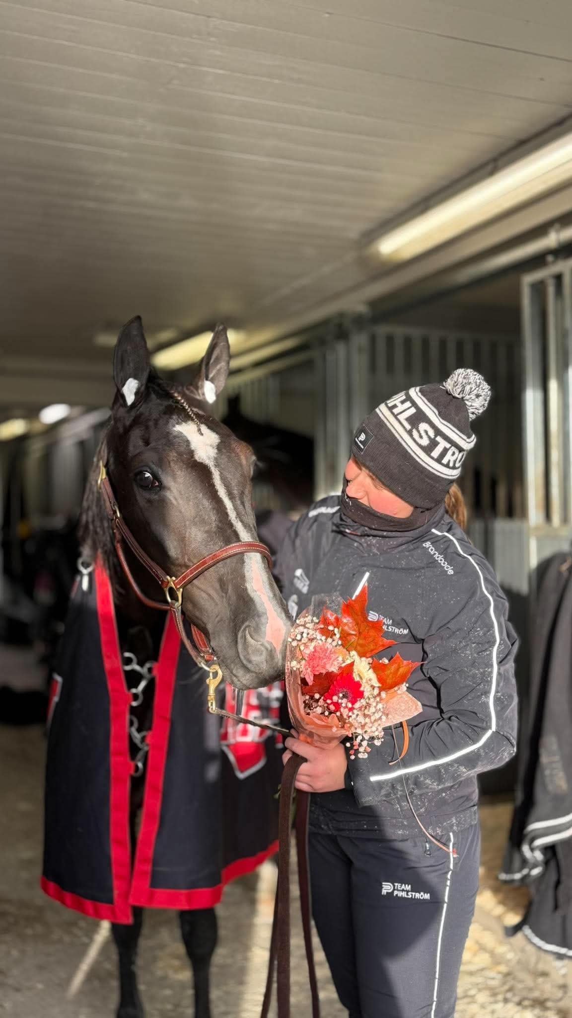 Person in black winter attire holding a black horse wearing a red blanket in a stable