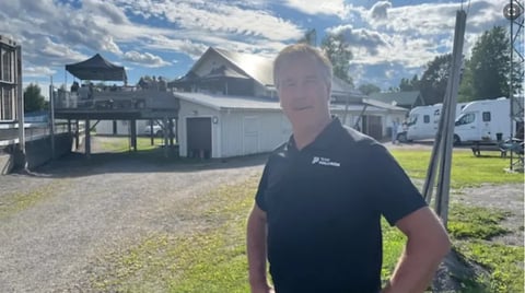 Man in black t-shirt standing outdoors at a farm or ranch with barns and mountains in background