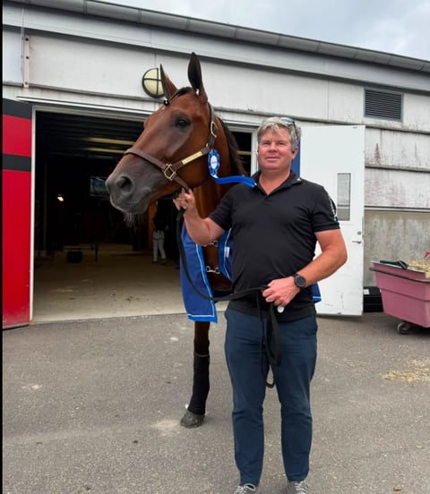 Man in black shirt standing with brown horse wearing blue blanket outside stable building